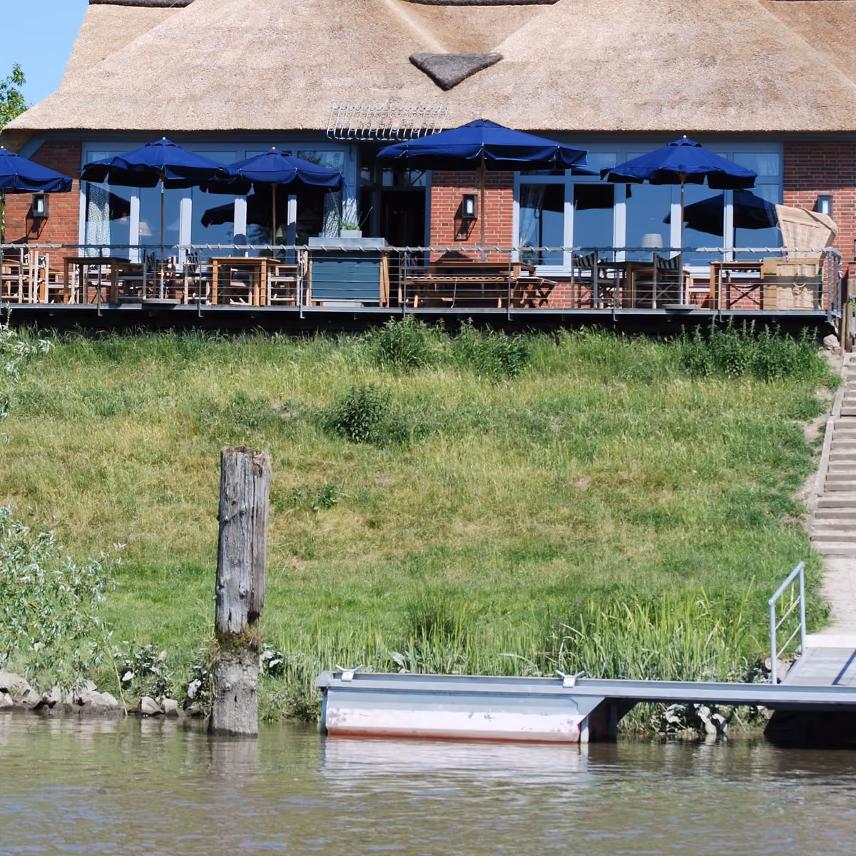 Der Schwimmsteg mit Gangway und einem festgemachten Boot, dahinter der Deich mit den Stufen hinauf zur Terrasse