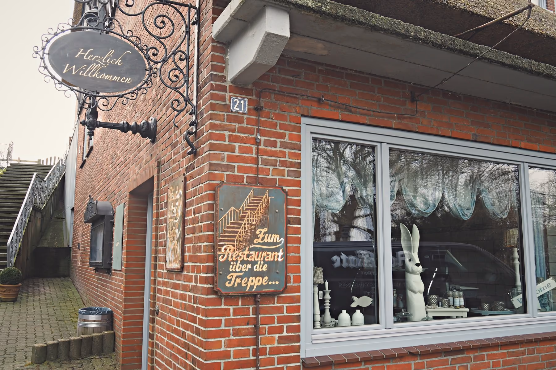 Der Eingang der Aal-Kate: unter dem Schild „Herzlich Willkommen" führt links eine Treppe mit Eisengeländer hinauf, an der Backsteinwand hängt ein gemaltes Schild mit der Aufschrift „Zum Restaurant über die Treppe"